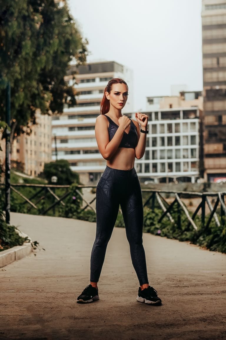 Full length of fit young female wearing sportive clothes and standing on road in city park with fists up and looking at camera